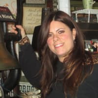 <p>Donna Massaro standing in front of a historic bell that was used for the Old Putnam Line at the Freight House Cafe.</p>