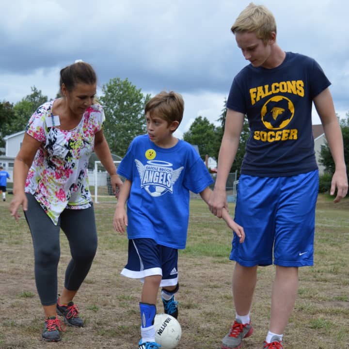 A young athlete with special needs dribbles up the field with his mother and a Saddle Brook Angels volunteer.