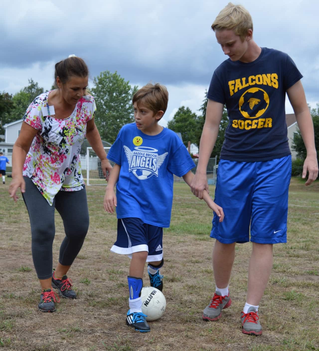 A young athlete with special needs dribbles up the field with his mother and a Saddle Brook Angels volunteer.