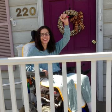 Christine Danza jubilantly raises one arm in front of her home. After breaking her neck, she is working hard to regain movement in her body but still cannot stand.