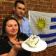 Sandra Salazar holds a chaja cake, a traditional Uruguayan pastry, while her husband Omar Gomez looks. The Uruguayan flag is in the background. The couple have opened Capri's Cuisine, specializing in Uruguayan pastries, in Norwalk.