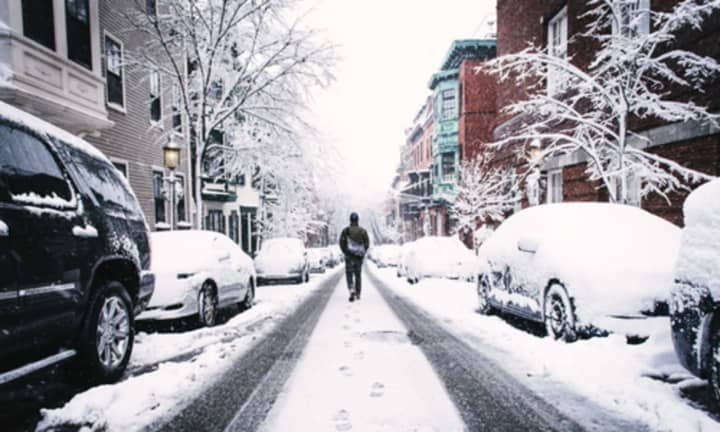 A person walks down a road flanked by snow-covered cars.