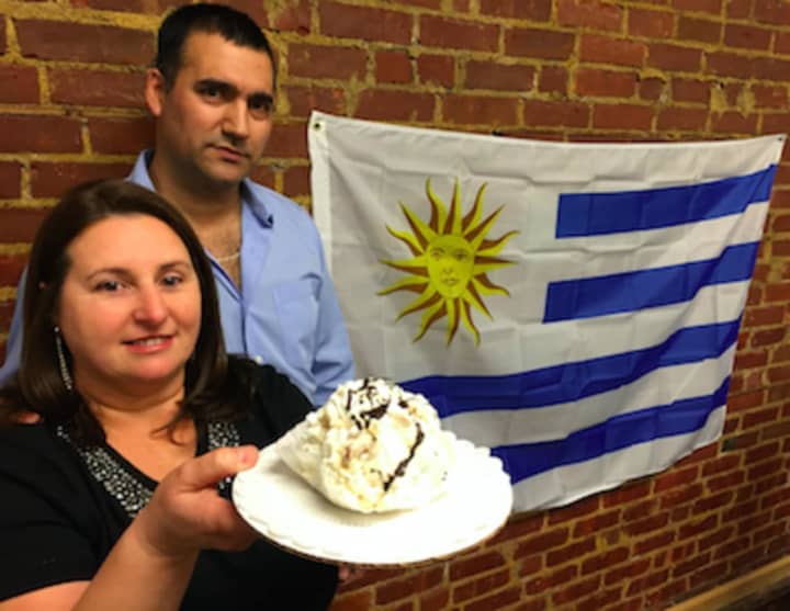 Sandra Salazar holds a chaja cake, a traditional Uruguayan pastry, while her husband Omar Gomez looks. The Uruguayan flag is in the background. The couple have opened Capri&#x27;s Cuisine, specializing in Uruguayan pastries, in Norwalk.