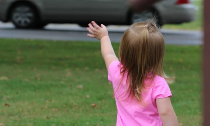 A young girl in a pink t-shirt waving at passing car.