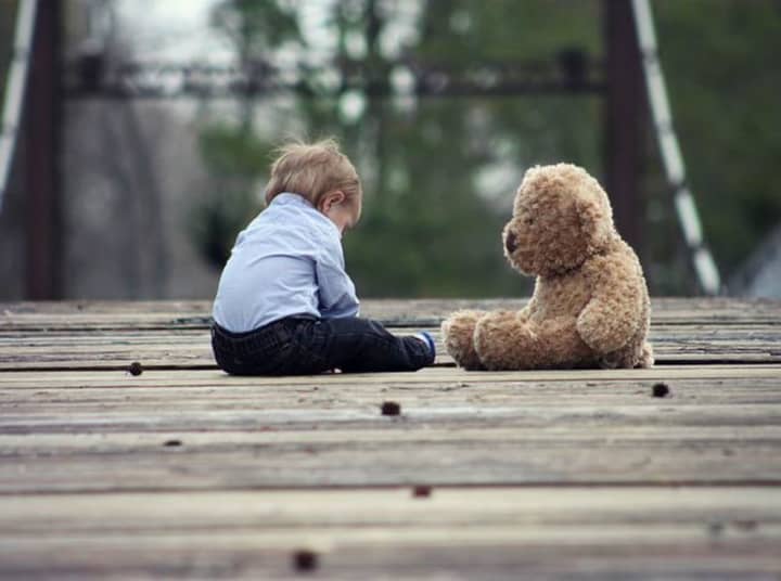A stock image of a young boy in a blue shirt sitting outside with a teddy bear.