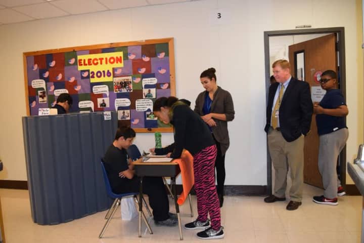 A student votes behind the privacy barrier while another signs in to vote. Teaching Assistant Ana Pascarelli and BOCES Superintendent James Ryan look on.